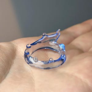 May include: A clear, blue-accented ring with a unique, fluid design. The ring features a series of small, rounded embellishments and a wave-like top. The ring is held in a hand, showcasing its delicate details and artistic form.