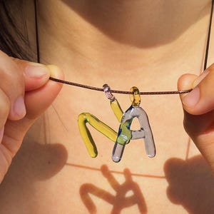 Puede incluir: Un collar con colgante de cristal hecho a mano que deletrea "NA". Las letras son de cristal amarillo transparente y gris azulado, suspendidas de un cord&oacute;n marr&oacute;n. El colgante est&aacute; sostenido por las manos de una persona.