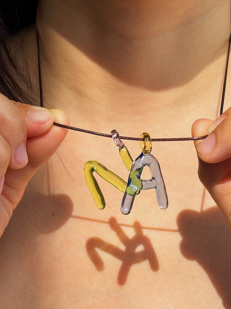 Puede incluir: Un collar con colgante de cristal hecho a mano que deletrea "NA". Las letras son de cristal amarillo transparente y gris azulado, suspendidas de un cord&oacute;n marr&oacute;n. El colgante est&aacute; sostenido por las manos de una persona.