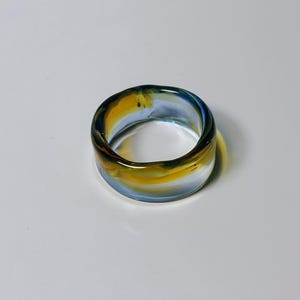 May include: A wide, clear glass ring with a marbled effect. The ring's band is a blend of yellow, blue, and brown. The ring is approximately 2.5 cm wide and is displayed on a white surface.