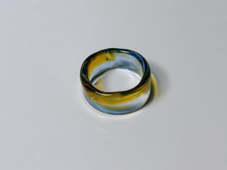 May include: A wide, clear glass ring with a marbled effect. The ring's band is a blend of yellow, blue, and brown. The ring is approximately 2.5 cm wide and is displayed on a white surface.