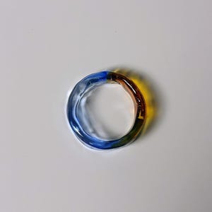 May include: A multi-coloured glass ring. The ring is circular and features a gradient of colours, including blue, yellow, green, and brown. The ring is made of transparent glass and is likely a piece of jewellery.
