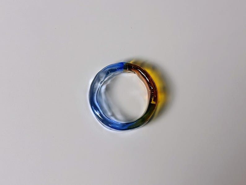 May include: A multi-coloured glass ring. The ring is circular and features a gradient of colours, including blue, yellow, green, and brown. The ring is made of transparent glass and is likely a piece of jewellery.