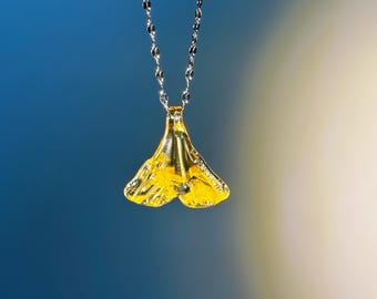 May include: A silver chain necklace featuring a yellow ginkgo leaf pendant. The pendant has a textured surface and hangs from a delicate chain. The background is a gradient of blue and white.