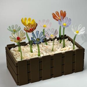 May include: A brown wooden planter box filled with white pebbles, showcasing colorful glass flowers. The flowers have various colors, including yellow, orange, blue, pink, and white, with green stems and yellow or red centers.