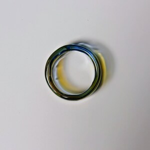 May include: A close-up of a ring with a distinctive design. The ring has a swirling, layered appearance with a mix of colours, including blue, green, and gold. The ring is made of a shiny, reflective material.