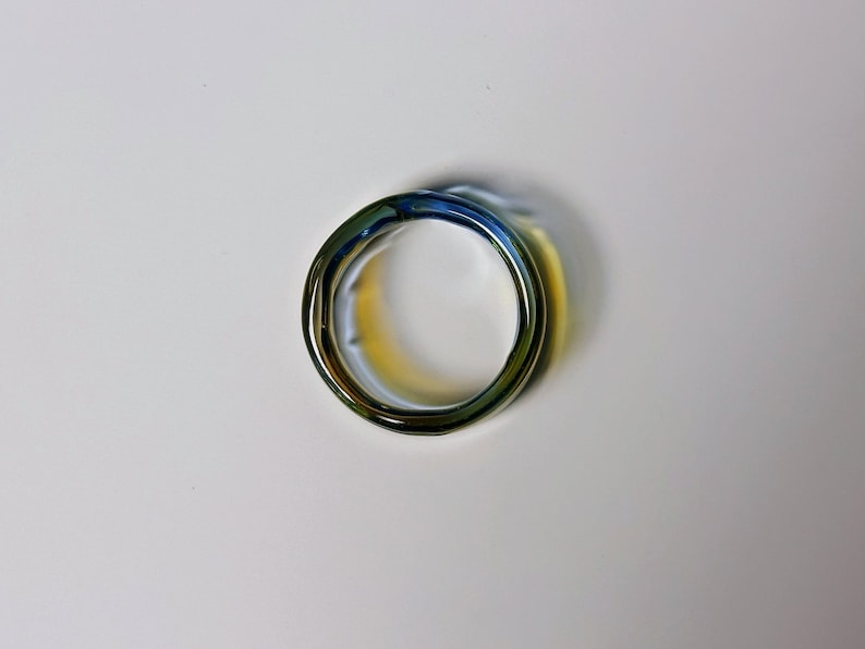 May include: A close-up of a ring with a distinctive design. The ring has a swirling, layered appearance with a mix of colours, including blue, green, and gold. The ring is made of a shiny, reflective material.