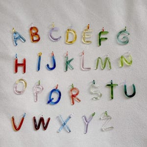 Puede incluir: Un conjunto de colgantes de letras del alfabeto de cristal de colores. Cada letra, de la A a la Z, es de un color diferente, incluyendo azul, naranja, morado, amarillo, verde y rojo. Las letras est&aacute;n suspendidas por peque&ntilde;os bucles met&aacute;licos.