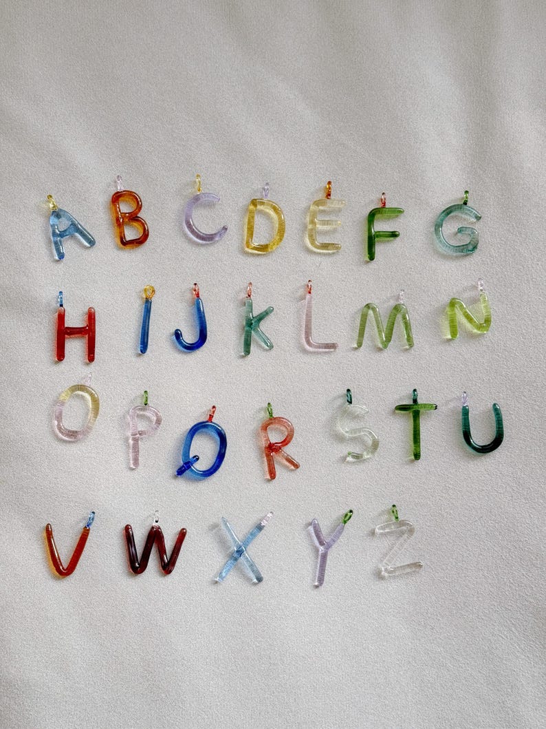 Puede incluir: Un conjunto de colgantes de letras del alfabeto de cristal de colores. Cada letra, de la A a la Z, es de un color diferente, incluyendo azul, naranja, morado, amarillo, verde y rojo. Las letras est&aacute;n suspendidas por peque&ntilde;os bucles met&aacute;licos.