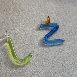 Puede incluir: Dos colgantes de cristal, uno con forma de la letra "J" en verde claro y otro con forma de la letra "Z" en azul. El colgante "Z" tiene un peque&ntilde;o lazo naranja en la parte superior. Ambos est&aacute;n sobre una superficie gris.