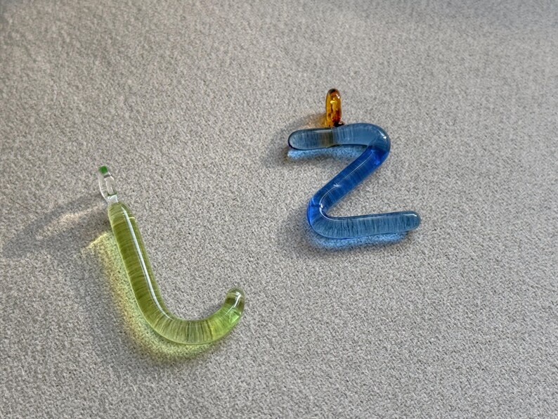 Puede incluir: Dos colgantes de cristal, uno con forma de la letra "J" en verde claro y otro con forma de la letra "Z" en azul. El colgante "Z" tiene un peque&ntilde;o lazo naranja en la parte superior. Ambos est&aacute;n sobre una superficie gris.