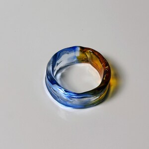 May include: A colourful glass ring, blending blue, yellow, and amber hues. This handcrafted ring is a unique piece of jewellery. The ring's design makes it a striking accessory, suitable for everyday wear or special occasions.