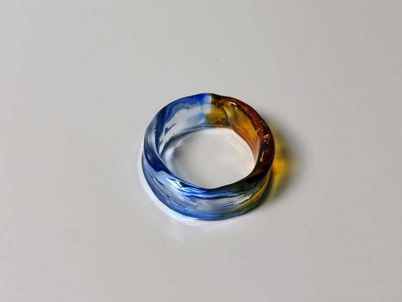May include: A colourful glass ring, blending blue, yellow, and amber hues. This handcrafted ring is a unique piece of jewellery. The ring's design makes it a striking accessory, suitable for everyday wear or special occasions.