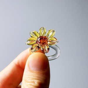 May include: A clear glass ring with a flower design. The flower has yellow petals and a brown center. The ring is held by a person's fingers against a gray background.