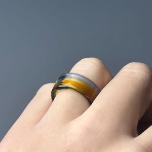 May include: A colourful ring with a band of yellow, orange, and black hues. The ring is worn on a finger, showcasing its unique design. The ring's colours create a striking contrast against the skin.