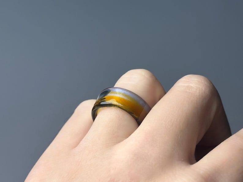 May include: A colourful ring with a band of yellow, orange, and black hues. The ring is worn on a finger, showcasing its unique design. The ring's colours create a striking contrast against the skin.