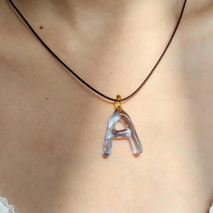 Puede incluir: Un colgante transparente con la letra "A" cuelga de un collar de cord&oacute;n negro. El colgante est&aacute; sujeto a una fianza dorada. El collar se muestra sobre un fondo neutro.