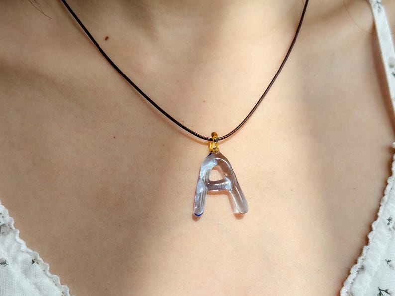 Puede incluir: Un colgante transparente con la letra "A" cuelga de un collar de cord&oacute;n negro. El colgante est&aacute; sujeto a una fianza dorada. El collar se muestra sobre un fondo neutro.