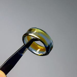 May include: A handcrafted glass ring with a distinctive design. The ring showcases a mix of yellow, blue, and clear hues, producing a captivating visual effect. The ring is held by black tweezers against a grey backdrop.