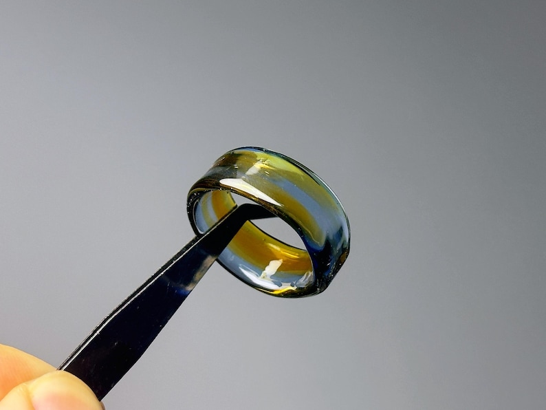 May include: A handcrafted glass ring with a distinctive design. The ring showcases a mix of yellow, blue, and clear hues, producing a captivating visual effect. The ring is held by black tweezers against a grey backdrop.