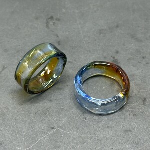 May include: Two glass rings with a colourful, iridescent finish. One ring has a yellow and green hue, while the other is blue and orange. The rings are thick and appear to be handmade.