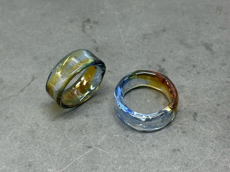 May include: Two glass rings with a colourful, iridescent finish. One ring has a yellow and green hue, while the other is blue and orange. The rings are thick and appear to be handmade.