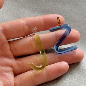 Puede incluir: Dos colgantes de cristal con forma de las letras "J" y "Z". La "J" es un degradado amarillo-verde, la "Z" es azul con detalles morados. Ambos tienen peque&ntilde;os bucles para colgar.