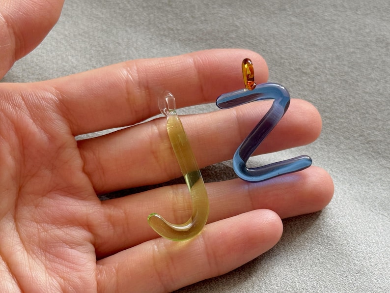 Puede incluir: Dos colgantes de cristal con forma de las letras "J" y "Z". La "J" es un degradado amarillo-verde, la "Z" es azul con detalles morados. Ambos tienen peque&ntilde;os bucles para colgar.