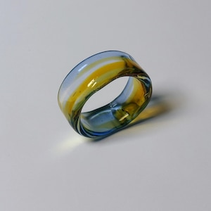 May include: A wide band glass ring with a swirl of colours. The ring's design incorporates blue, yellow, and white. A unique piece of jewellery.