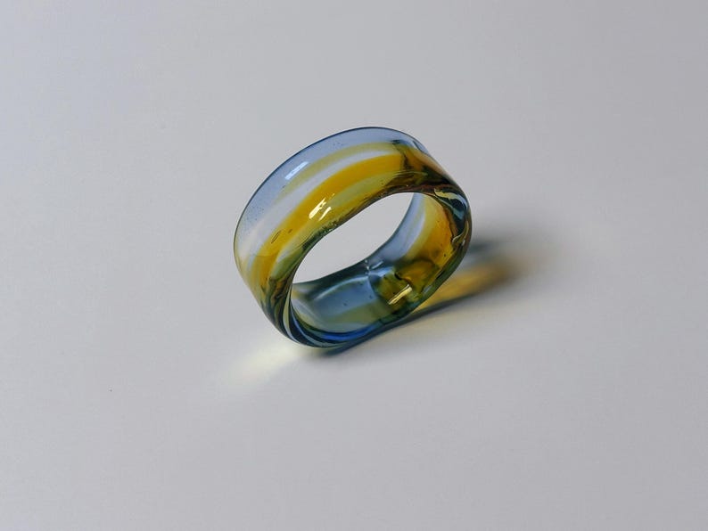 May include: A wide band glass ring with a swirl of colours. The ring's design incorporates blue, yellow, and white. A unique piece of jewellery.