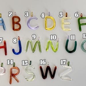 Puede incluir: Una colecci&oacute;n de colgantes de letras de vidrio de colores, cada uno con un peque&ntilde;o lazo para colgar. Las letras son de varios colores, incluyendo azul, naranja, verde y amarillo. Las letras est&aacute;n dispuestas sobre un fondo gris claro.