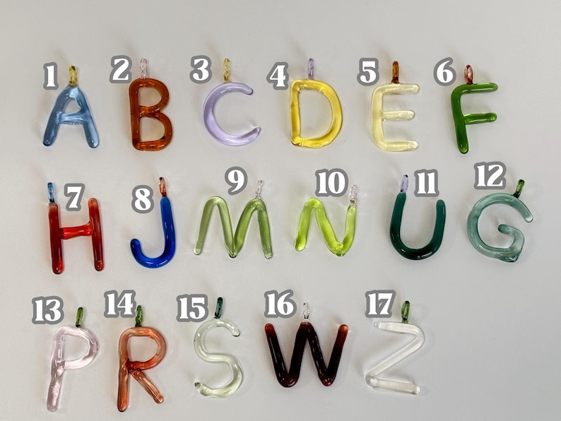 Puede incluir: Una colecci&oacute;n de colgantes de letras de vidrio de colores, cada uno con un peque&ntilde;o lazo para colgar. Las letras son de varios colores, incluyendo azul, naranja, verde y amarillo. Las letras est&aacute;n dispuestas sobre un fondo gris claro.