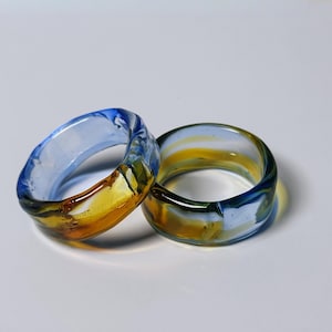 May include: Two glass rings with a marbled design. The rings feature a combination of blue, yellow, and clear glass. The rings are thick and appear to be handmade. The rings are displayed on a white surface.