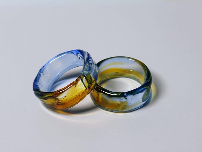 May include: Two glass rings with a marbled design. The rings feature a combination of blue, yellow, and clear glass. The rings are thick and appear to be handmade. The rings are displayed on a white surface.