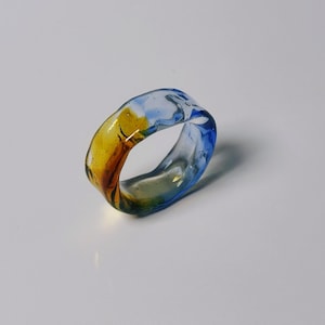 May include: A colourful glass ring with a marbled design. The ring features shades of blue, yellow, and brown. The transparent glass allows light to pass through, creating a unique visual effect. A stylish accessory.