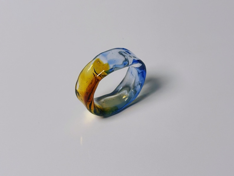 May include: A colourful glass ring with a marbled design. The ring features shades of blue, yellow, and brown. The transparent glass allows light to pass through, creating a unique visual effect. A stylish accessory.