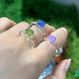 May include: Three clear glass rings with colorful, rounded tops. One ring has a green top, another a blue top, and the third a purple top. The rings are on a hand, with a green, mossy background.