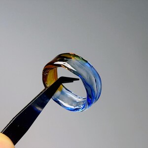 May include: A glass ring with a swirling design in shades of blue, yellow, and clear. The ring is held by black tweezers against a grey background, highlighting its transparent and artistic qualities.