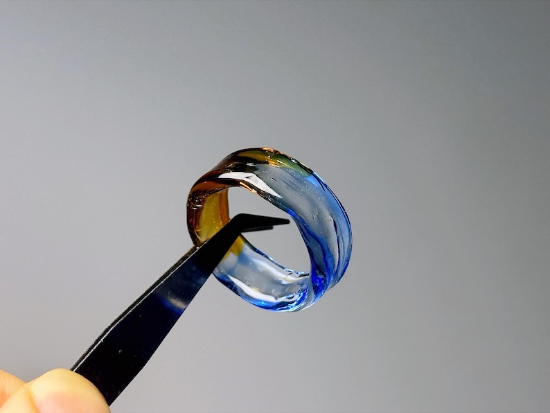 May include: A glass ring with a swirling design in shades of blue, yellow, and clear. The ring is held by black tweezers against a grey background, highlighting its transparent and artistic qualities.
