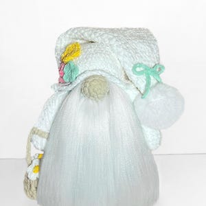 May include: A handmade gnome doll with a long white beard and a white hat adorned with flowers and a pom-pom. The gnome is holding a small basket with a daisy design. The doll is made of soft, textured materials.