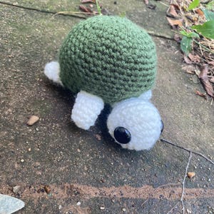 Op de afbeelding: Een handgemaakt gehaakt schildpad speelgoed. De schildpad heeft een groene schild, witte kop en ledematen en zwarte ogen. Het speelgoed ligt op een betonnen oppervlak.