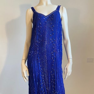 May include: A long, royal blue dress with a V-neck and thin straps. The dress is adorned with silver beading in a feather design. The hemline features a tiered, ruffled detail. The dress is displayed on a mannequin.