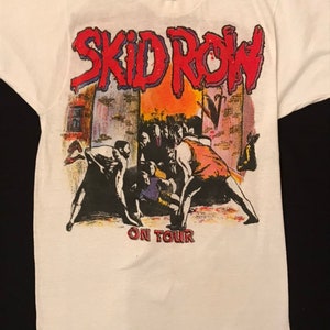 Op de afbeelding: Wit t-shirt met de bandnaam "SKID ROW" in rood, omlijnd in blauw, boven een afbeelding van een menigte en figuren. De tekst "ON TOUR" staat onder de afbeelding. Het ontwerp heeft een vintage, concert-achtige uitstraling.