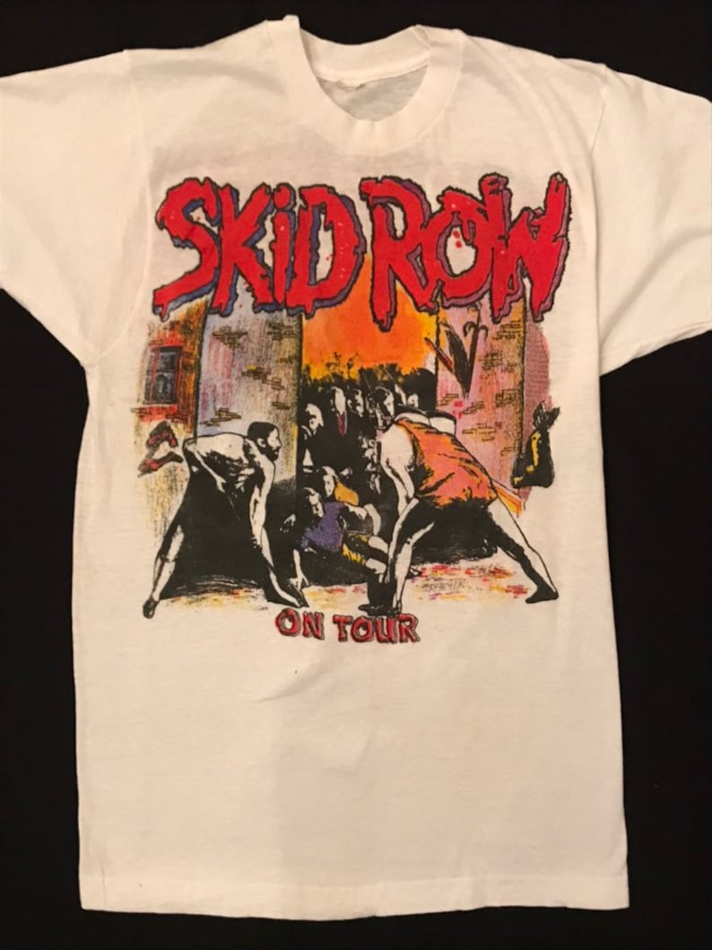 Op de afbeelding: Wit t-shirt met de bandnaam "SKID ROW" in rood, omlijnd in blauw, boven een afbeelding van een menigte en figuren. De tekst "ON TOUR" staat onder de afbeelding. Het ontwerp heeft een vintage, concert-achtige uitstraling.