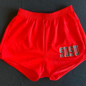 Può includere: Pantaloncini sportivi rosso vivo con elastico in vita. I pantaloncini presentano le lettere "SDSU" in nero con contorno bianco. Questi pantaloncini sono probabilmente vintage e adatti alle attività sportive.