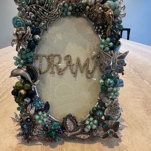 May include: Ornate oval picture frame with the word "DRAMA" in gold glitter. The frame is heavily embellished with turquoise, green, and silver-toned beads, flowers, and butterfly accents. The frame is a decorative item.
