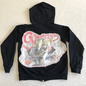 Può includere: Felpa con cappuccio nera con zip, con una grafica 'Grease' bianca e rosa sul retro. La grafica presenta un'immagine di due persone in stile cartone animato.