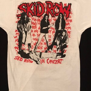 Op de afbeelding: Wit t-shirt met een afbeelding van de band Skid Row in concert. Het ontwerp bevat de naam van de band in het rood, samen met zwart-wit illustraties van de bandleden. De woorden "IN CONCERT" staan ook in het rood.