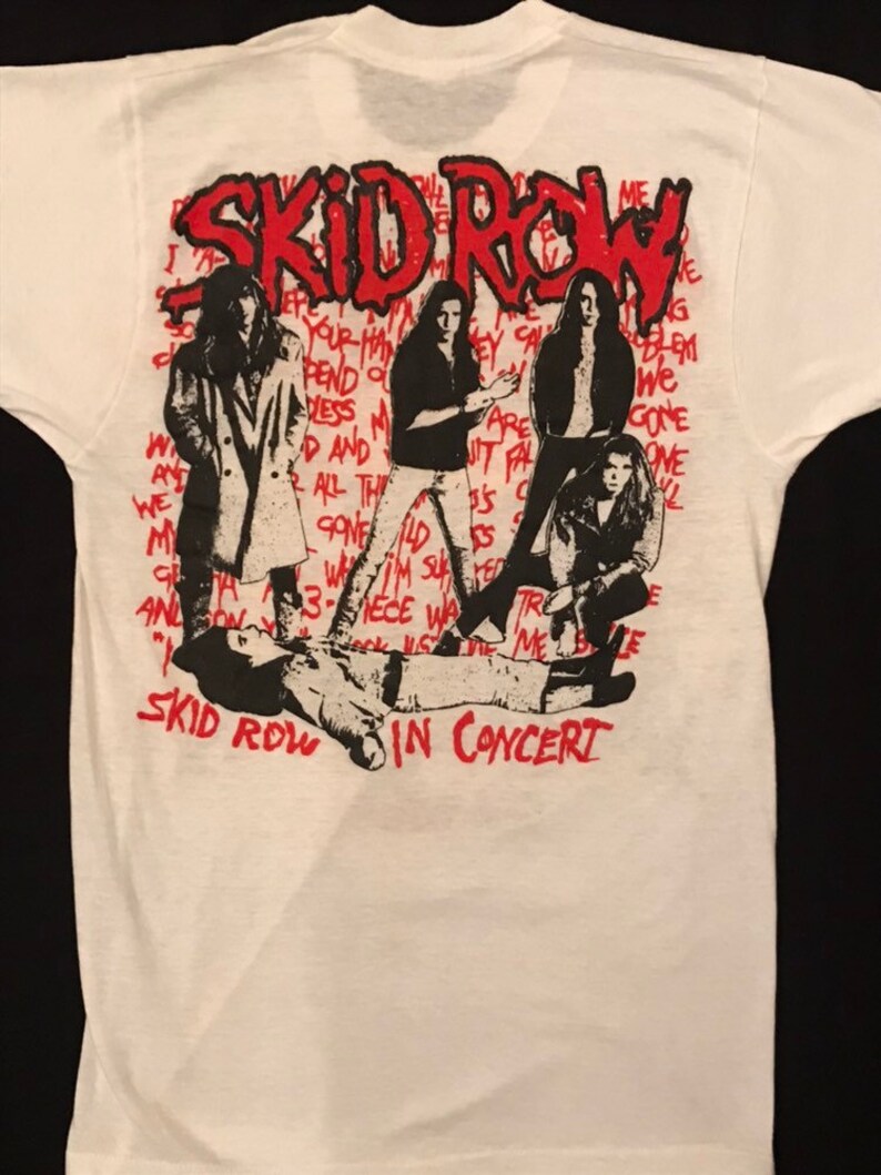 Op de afbeelding: Wit t-shirt met een afbeelding van de band Skid Row in concert. Het ontwerp bevat de naam van de band in het rood, samen met zwart-wit illustraties van de bandleden. De woorden "IN CONCERT" staan ook in het rood.