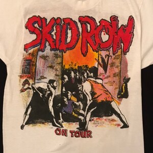 Op de afbeelding: Wit T-shirt met de bandnaam "SKID ROW" in rood en blauw, boven een afbeelding van een menigte. De tekst "ON TOUR" staat eronder in het rood. Het T-shirt is gemaakt van zacht, wit materiaal en is geschikt voor dagelijks gebruik.